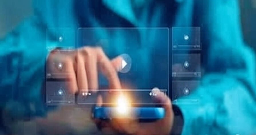 A person in a blue shirt interacting with a holographic interface, pressing a play button on a video player displayed above a glowing smartphone held in their hands. The interface includes multiple video thumbnails with play icons, set against a blue background.